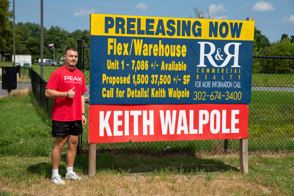 Cameron Horn of PEAK Fitness standing beside a Keith Walpole Group "For Lease" sign outside the warehouse units at 26850 Lewes Georgetown Highway in Harbeson, Delaware.
